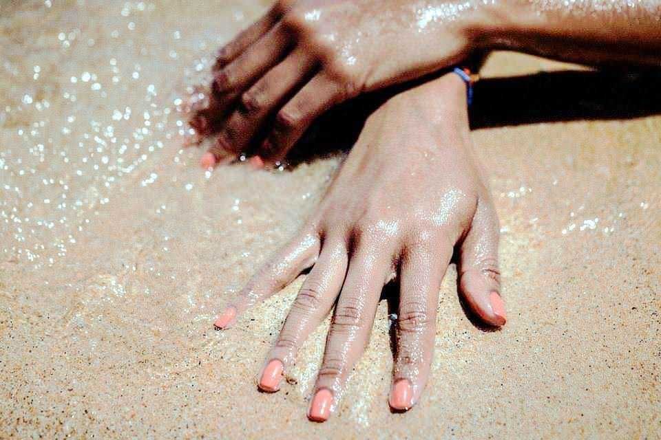 Mani mani con unghie curate e smalto rosa su spiaggia di sabbia dorata sotto la luce del sole, scena di relax e divertimento estivo.