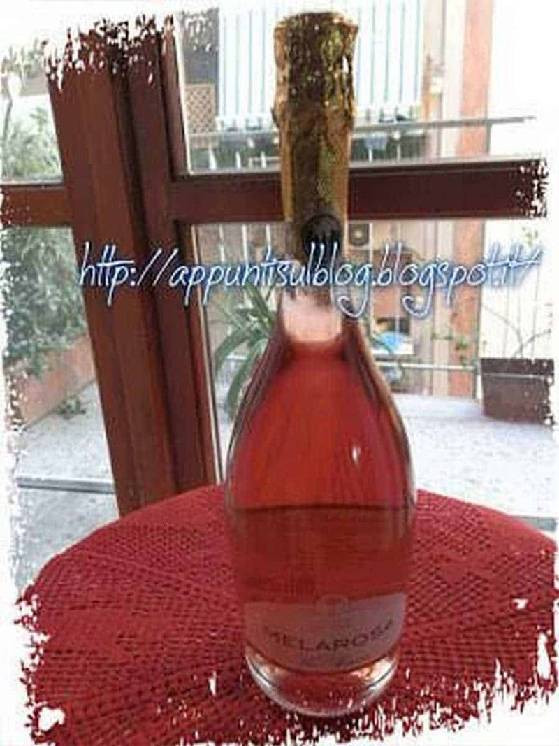 Cantine 2 Palme, vini di qualità fruttati dal sapore delicato 2 Vini del salento: Bottiglia di vino rosso con cappuccio decorato, appoggiata su tovaglia rossa, in un ambiente domestico con finestra e balcone sullo sfondo. Perfetta per foto di degustazioni e momenti conviviali.