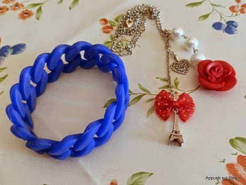 Colorato braccialetto in plastica blu, collana con pendente a forma di rosa rossa, farfalla rossa e torre Eiffel, su tovaglia con fiori. Accessori di necklaces e bracciali moda, stile femminile e trendy.