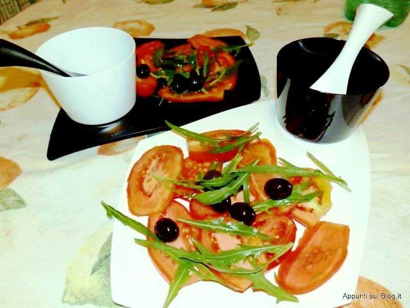 Pomodori pelati in scatola, conserve di olive nere denocciolate, sottaceti di cipolle e peperoncini, e snack di olives greche e sott'olio su tavolo con tovaglia floreale.