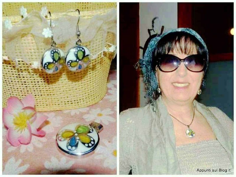 Orecchini e collana artigianali con farfalle colorate, esposte su un contenitore di vimini giallo e posizionate su un tovagliolo con fiori, accanto a una donna sorridente che indossa gli stessi gioielli, con occhiali scuri e sciarpa in testa.