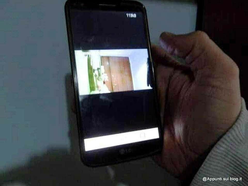Sfondo di una mano che tiene uno smartphone LG, mostrando un'immagine di un'entrata con porta in legno e dettaglio di arredamento moderno, scattato in condizioni di scarsa illuminazione.