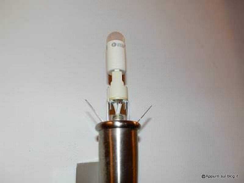 Lampada led impressa in un tubo di metallo, accesa, vicino alla parete bianca.