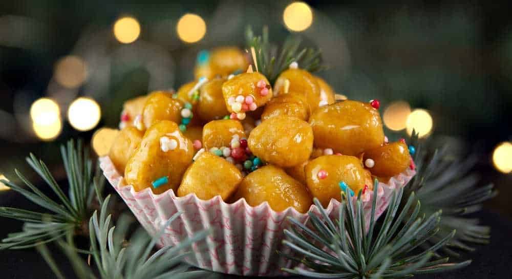 Baci di dama decorati con zuccherini colorati in una coppetta di carta natalizia, circondati da rami di pino, perfetti per le festività natalizie.