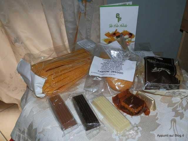 Filamento di pasta lunga come spaghetti avvolto in carta, vari cioccolatini e un pacchetto di dolciumi con etichetta, in un ambiente con tovaglia decorativa.