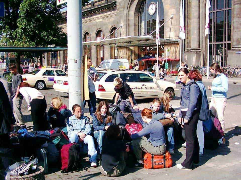 Viaggi gruppi o soli: Viaggiare in gruppo o da soli: Famiglia di viaggiatori con bagagli e valigie in attesa davanti a una stazione ferroviaria in Germania. Atmosfera di viaggio con persone pronte alla partenza, ambientazione urbana e mezzi di trasporto visibili.
