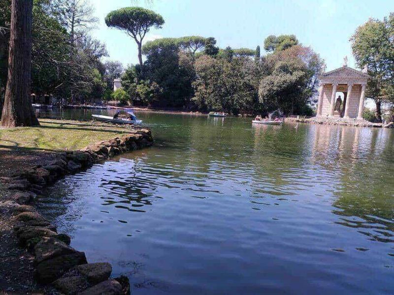 Roma capitale tour in mesi caldi 5 Tranquillo lago con barche a remi, circondato da grandi alberi e vegetazione lussureggiante in un parco cittadino, con un'architettura storica in stile neoclassico sullo sfondo.