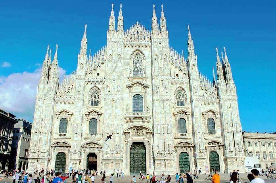 Maestosa cattedrale gotica Duomo di Milano con facciata in marmo, archi e vetrate artistiche, visitata da numerosi turisti in una giornata soleggiata.