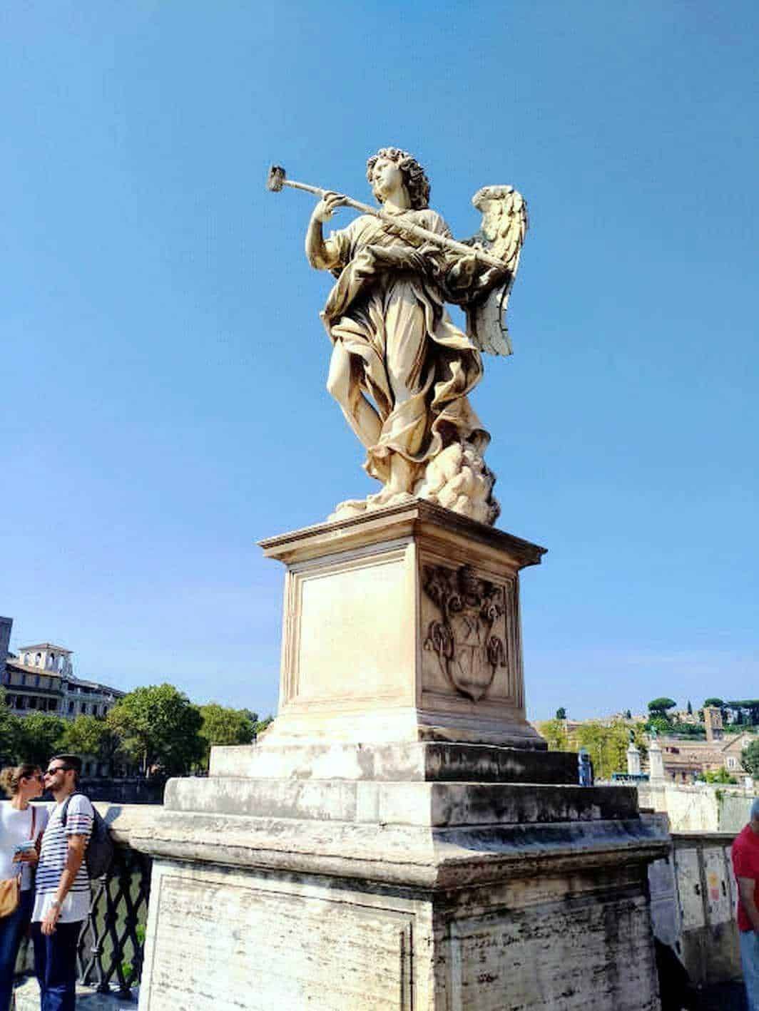 Statua di un angelo in pietra con tromba, cielo azzurro sullo sfondo, simbolo di arte e storia, situata in un luogo storico di Roma. Perfetta per articoli di turismo culturale e patrimonio artistico.