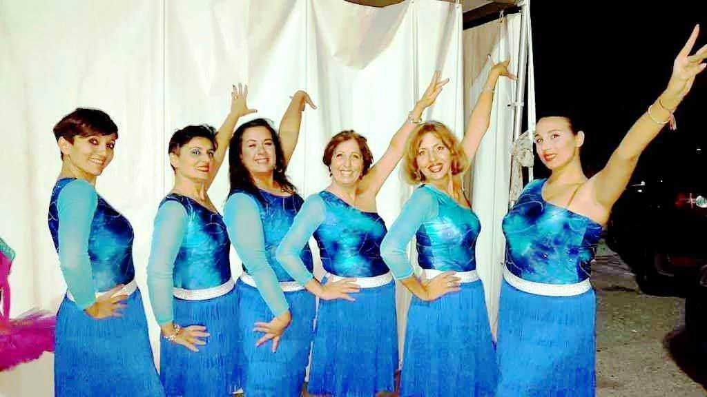 Elegante gruppo di donne in abiti coordinati blu con motivi celestiali, sorridenti e in pose di festa, pronto a esibirsi in uno spettacolo di danza o performace artistica, sul palco.