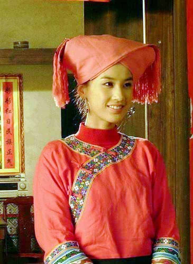 Elegante donna asiatica in abiti tradizionali cinesi con cappello rosso e dettagli ricamati, sorridente, in un ambiente domestico con decorazioni e arredi tipici.