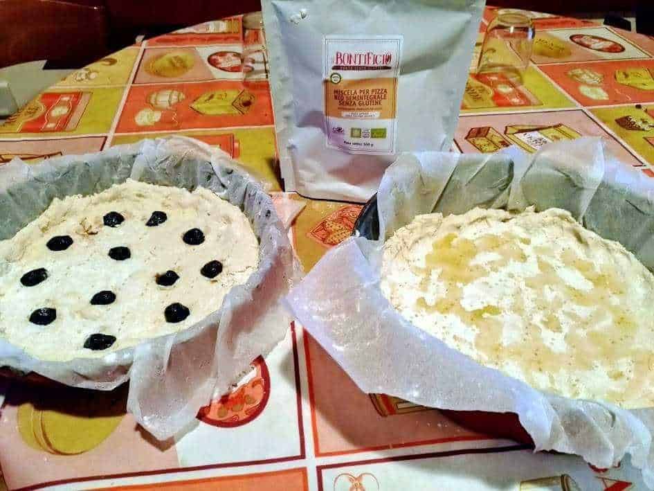 Focaccia senza glutine con olive nere, preparata in teglia, avvolta in carta forno, su tovagliolo allegro colorato, con un sacchetto di farina senza glutine sullo sfondo, ideale per cibi senza glutine, ricetta casalinga.