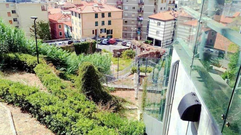 La Spezia siti d'interesse storici e moderni 8 Vista panoramica di un giardino urbano con piante e arbusti accanto a un edificio moderno con balcone in vetro, in un quartiere residenziale.