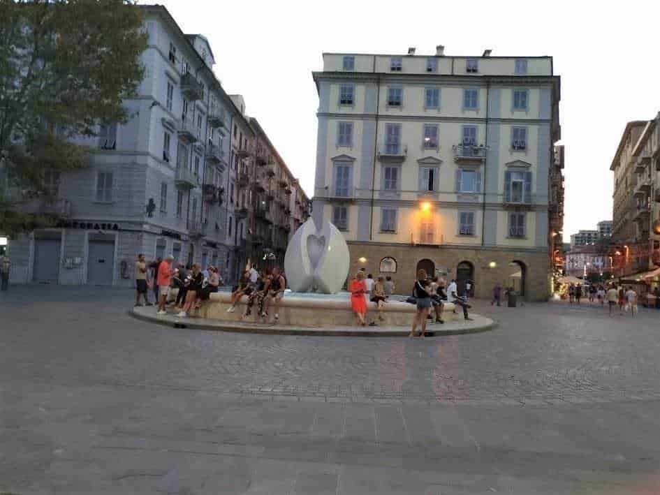 Fontana pubblica con sculture moderne in una piazza urbana a tarda sera, circondata da edifici storici e con persone che si rilassano sugli scalini, creando un’atmosfera vivace e accogliente.