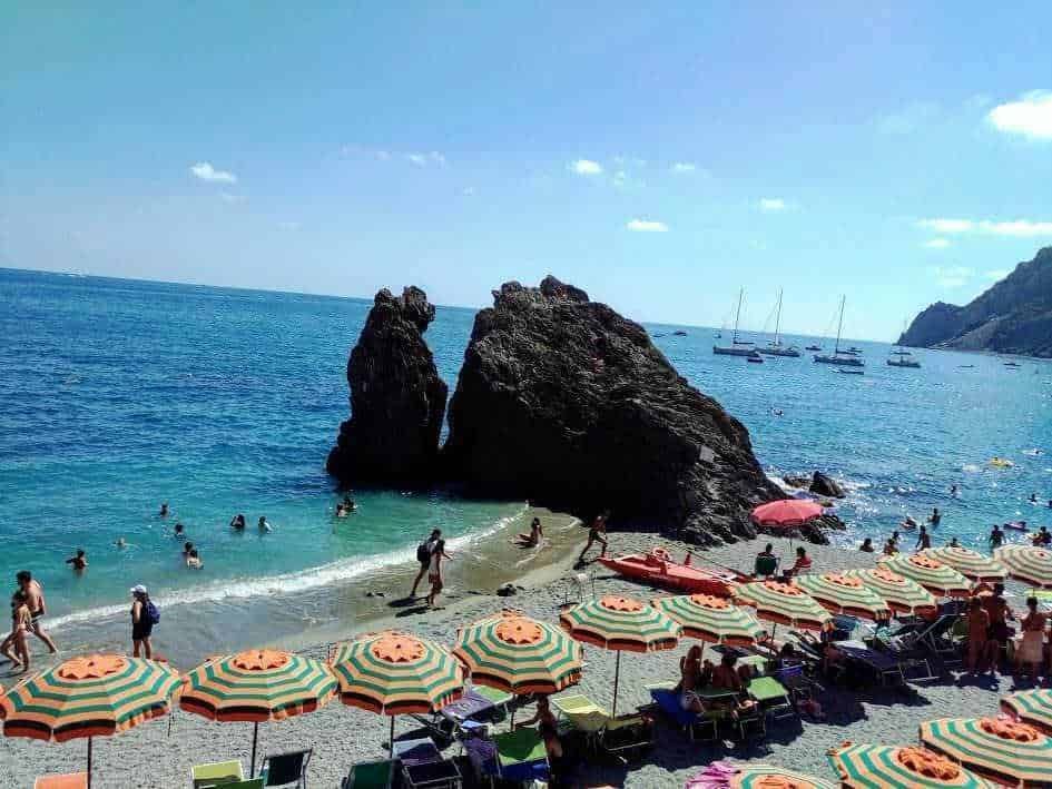 Splendida spiaggia con grandi scogli, lettini e ombrelloni colorati, persone che nuotano e si rilassano al sole, acque cristalline e baia pittoresca con barche a vela sullo sfondo.