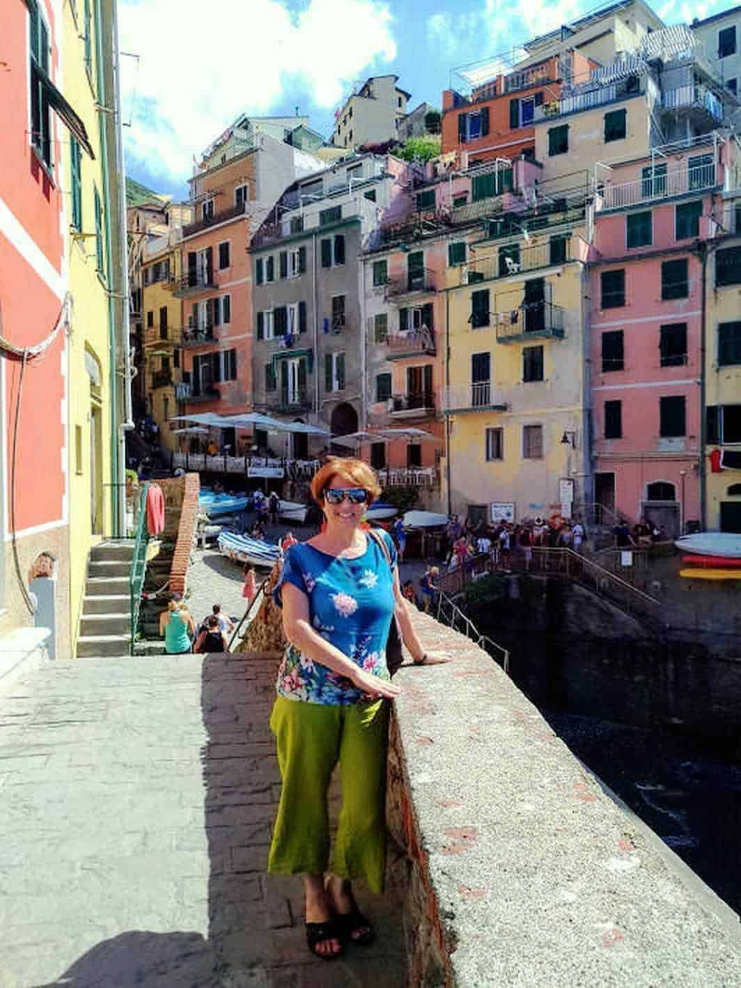 Colorate case di pescatori color pastello a Vernazza, Cinque Terre, Italia, con una donna sorridente in primo piano, abbigliamento estivo, atmosfera turistica e paesaggio marittimo.