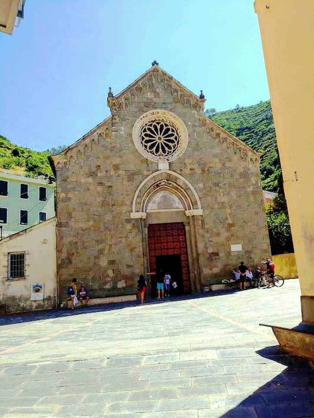 Chiesa storica con architettura semplice e dettagli rosetta, immersa in un centro abitato con persone e biciclette, circondata da colline verdi e un cielo azzurro, perfetta per articoli su turismo e cultura.