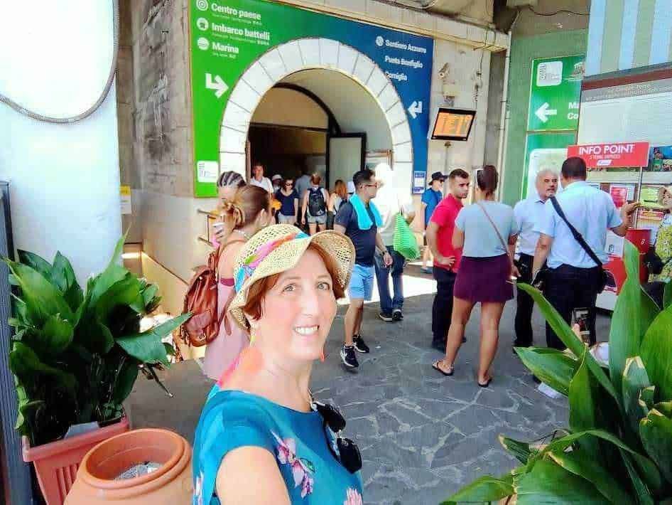 Stazione ferroviaria con molte persone in fila, segnaletica verde e ingresso architettonico, donna sorridente in primo piano, piante decorative sullo sfondo, viaggio e turismo, attesa dei treni, informazione e mobilità.