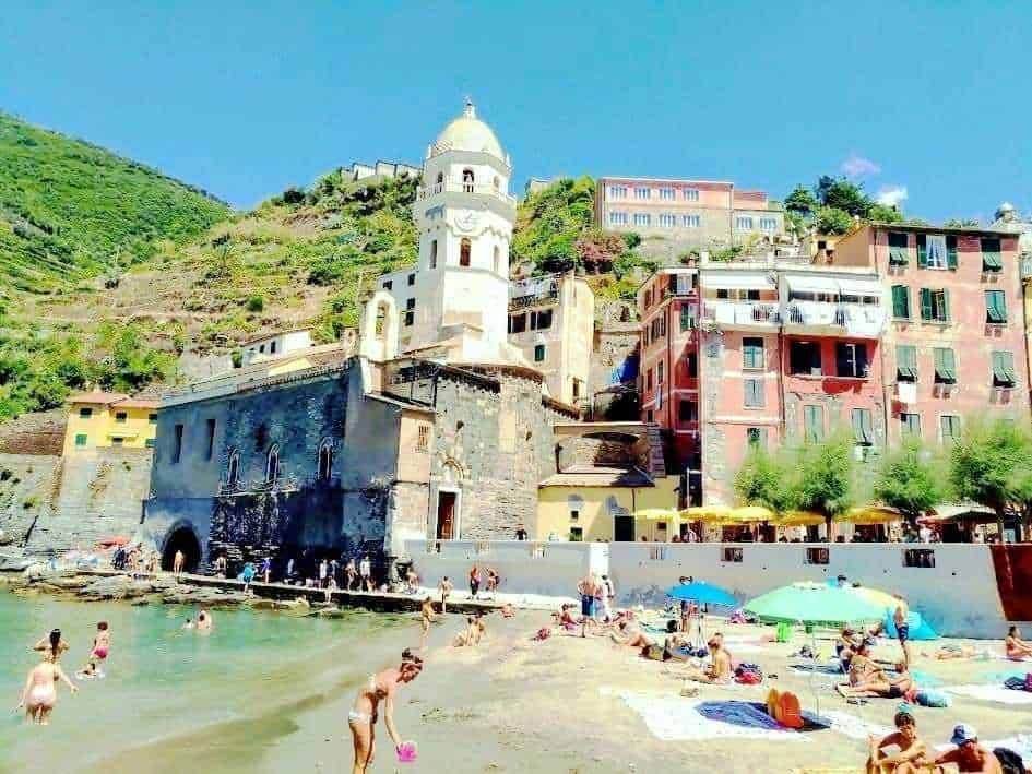 Appunti Sul Blog, spiaggia affollata con persone che nuotano e prendono il sole, vicino a edifici colorati e una chiesa storica sul mare, ideale per il turismo e le vacanze estive in Italia.
