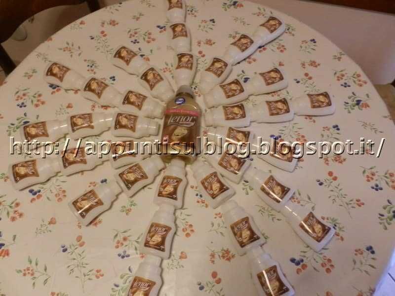 Bottiglie di vino disposte a raggiera su tavolo con tovaglia floreale, decorazione originale e creativa per feste o eventi speciali.