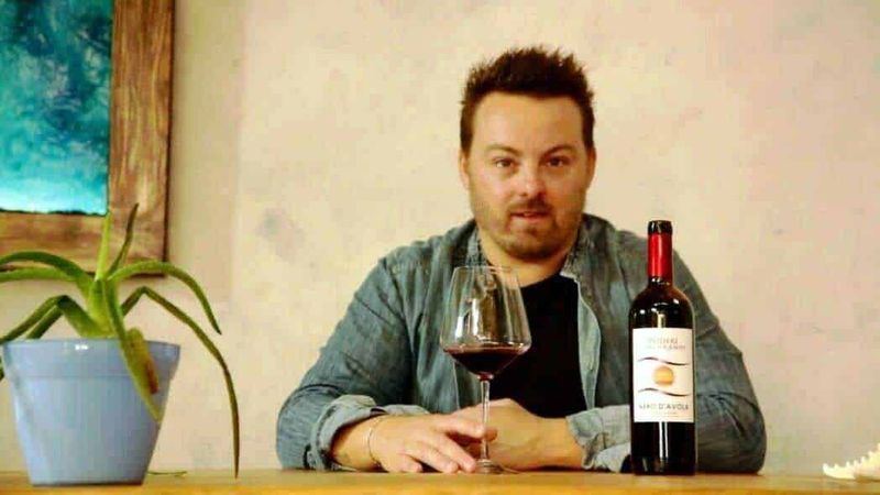 Linea di vini integralmente prodotti da Eurospin 2 Un uomo che beve vino rosso in un ambiente domestico con una bottiglia di vino, un bicchiere e una pianta sul tavolo, atmosfera accogliente e rilassante.