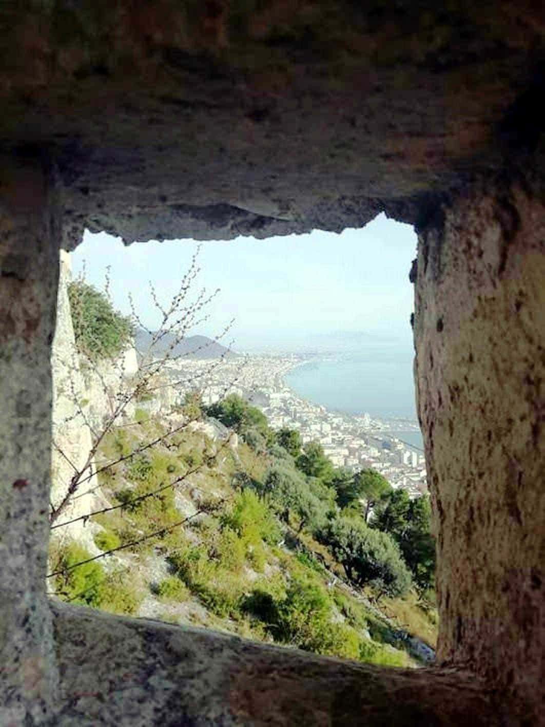 Veduta panoramica della costa e della città vista attraverso una fessura in una torre di pietra, con vegetazione verde e mare sullo sfondo, in un ambiente storico e naturale di grande fascino.