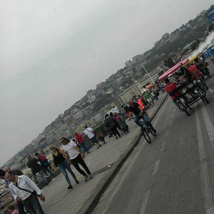 Pedalare in bicicletta: perché fa bene. Passeggiata sul lungomare di Napoli con pedoni, ciclisti e carri di cibo sotto un cielo nuvoloso, con la città sullo sfondo. 