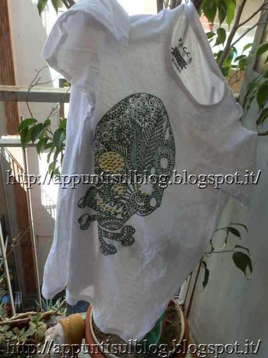 Una maglietta bianca con un design artistico di una testa di scheletro decorata con pattern intricati, appesa tra piante verdi in un ambiente naturale e luminoso.