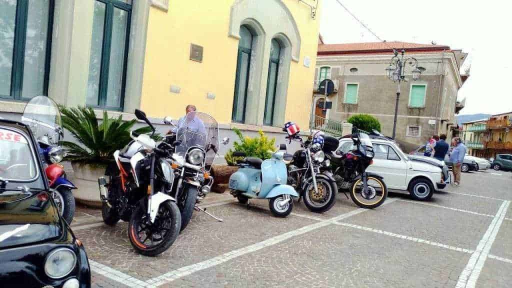 Moto, scooter e auto parcheggiate davanti a un edificio in un parcheggio pubblico, scena urbana con persone che si godono il giorno all'aperto, fotografia di veicoli e ambientazione cittadina.
