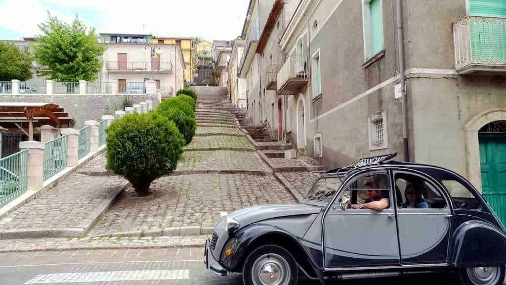 L'immagine mostra un'auto classica in stile Volkswagen Beetle parcheggiata su una strada in salita in un quartiere residenziale di città in Italia, con case vecchie e un vialetto stretta con un albero ben curato.