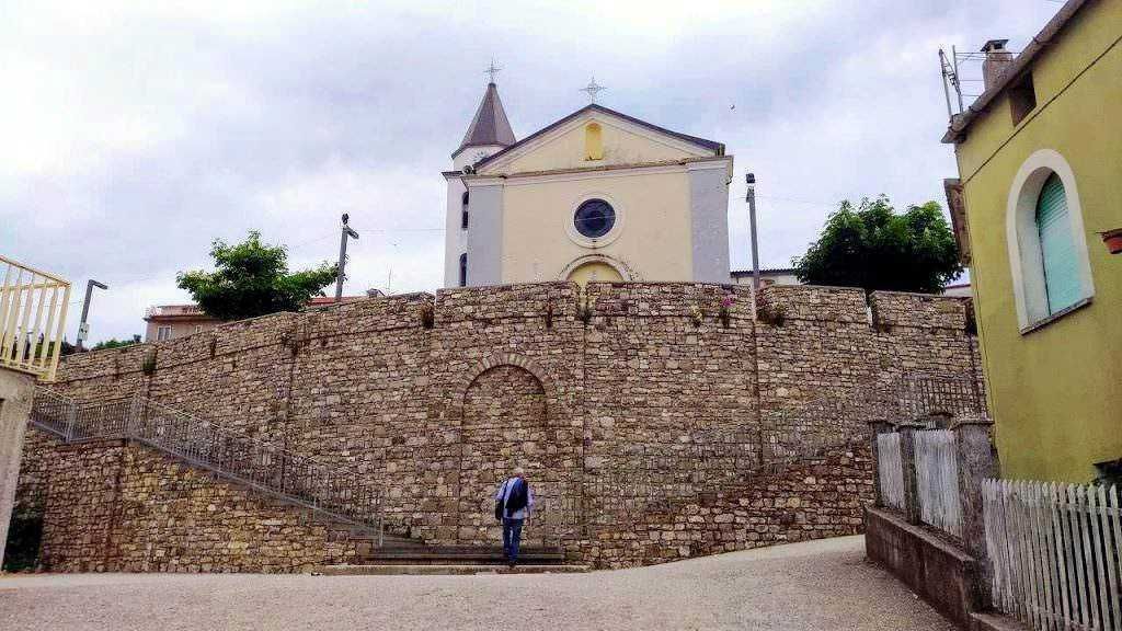 Steeple of a small church with a stone wall in an Italian village, surrounded by colorful houses and green trees, under a cloudy sky.