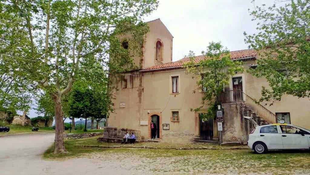Vecchia chiesa in pietra circondata da alberi e auto parcheggiate, esempio di architettura storica in un paesaggio rurale, ideale per appassionati di storia e cultura locale.