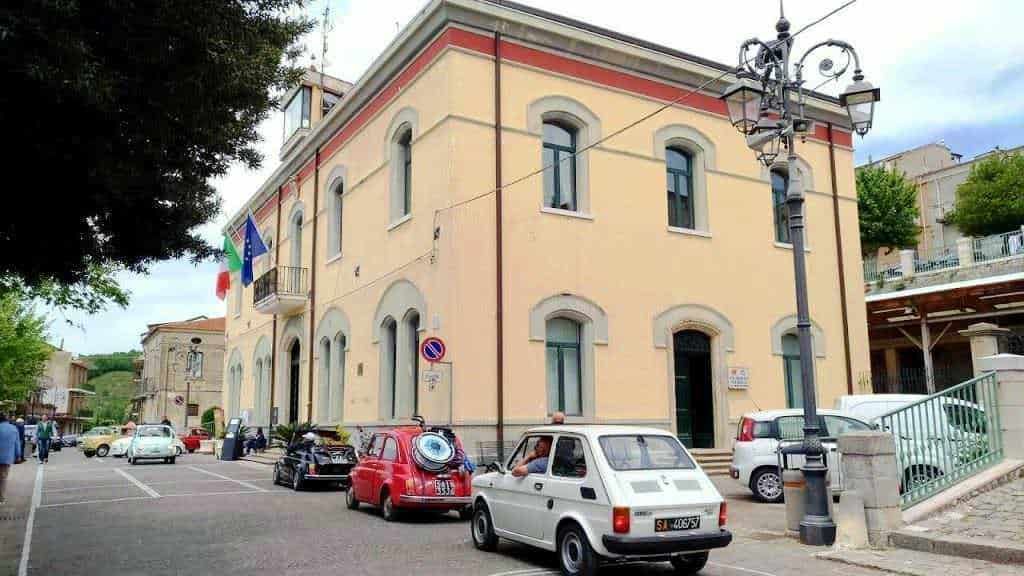 Auto storiche e edificio storico in un affascinante scorcio di centro città, con bandiere italiane ed europee, lampioni vintage e veicoli d'epoca che arricchiscono l'atmosfera culturale e storica del luogo.