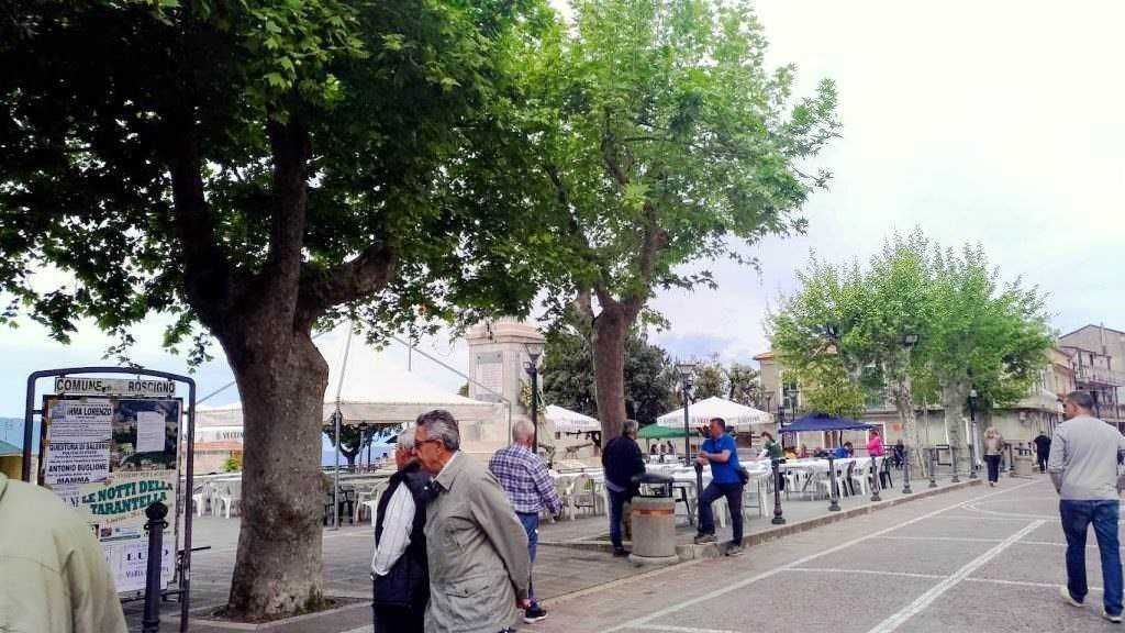 Passeggiata nel centro di Rossano, con albero al centro e persone che si godono la domenica in un'atmosfera tranquilla e vivace, tra negozi e panchine.