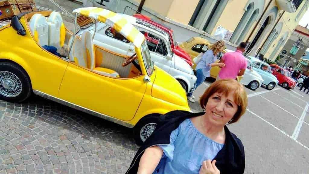 Fiat 500 cabriolet anni '70 di colore giallo esposta a un raduno di auto d'epoca con persone che ammirano i veicoli in un centro città.