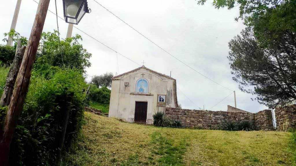 Chapella di campagna situata su una collina con muro in pietra, alberi e vegetazione circostante, cielo nuvoloso, strada sterrata in salita, ambiente rurale e tranquillo.