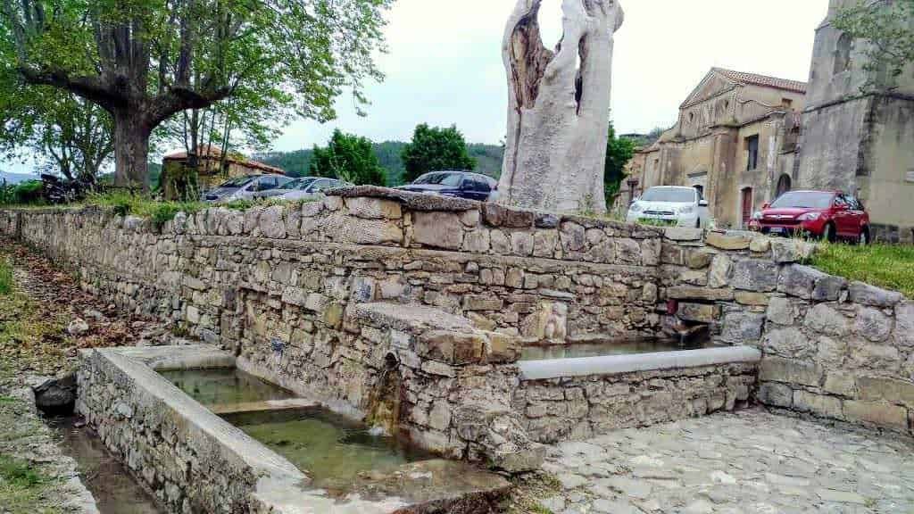 Fontana storica in pietra con acqua che scorre, situata in un contesto rurale con alberi e edifici antichi, ideale per appunti sul blog riguardo storia, architettura e paesaggi tradizionali italiani.