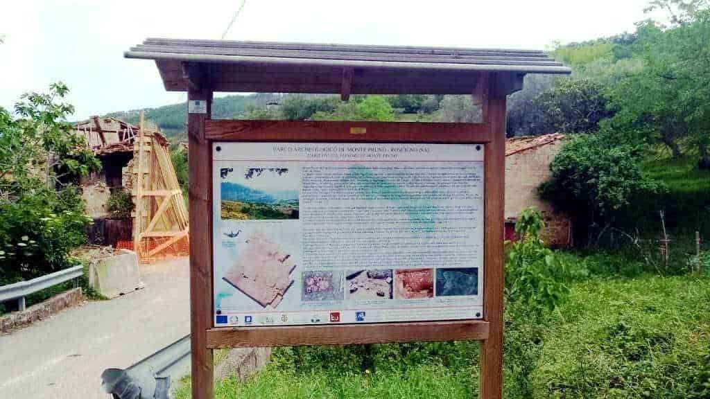 Pannello di info archeologica all'aperto in un contesto rurale, con testo e immagini illustrative, situato lungo la strada principale in un ambiente naturale con vegetazione e case tradizionali nelle vicinanze.