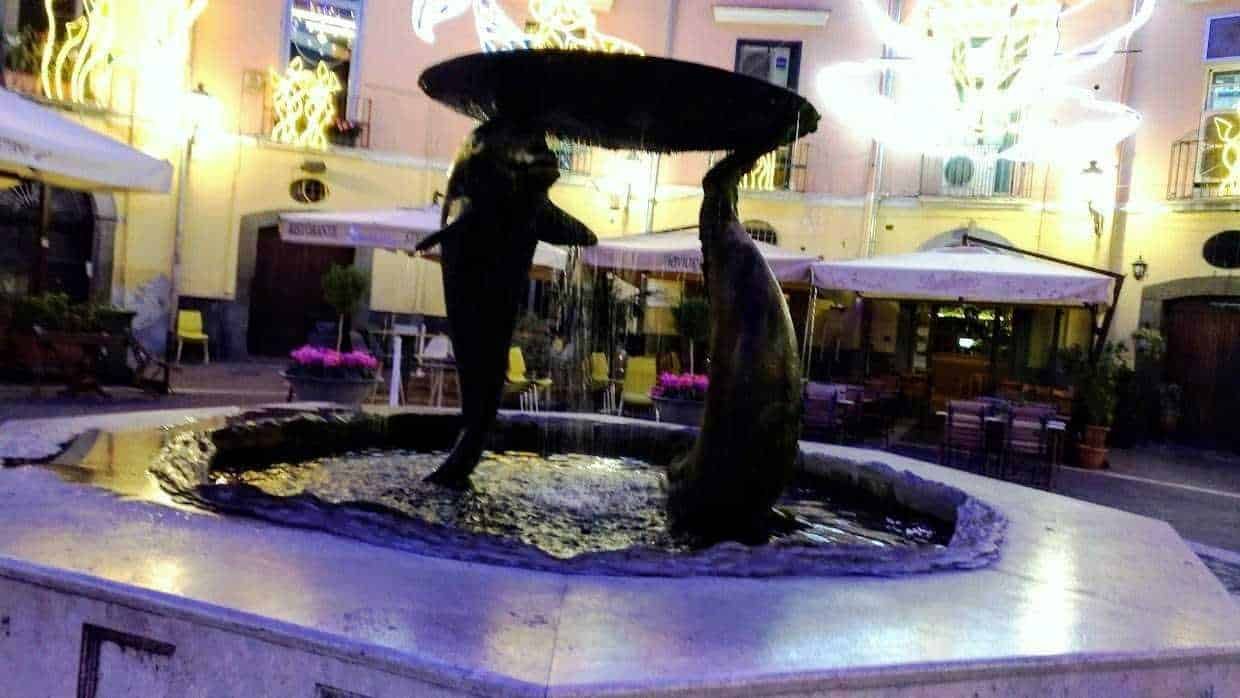 Antico fontana in piazza con sculture di delfini, illuminata di sera, attrazione storica e artistica in centro città.