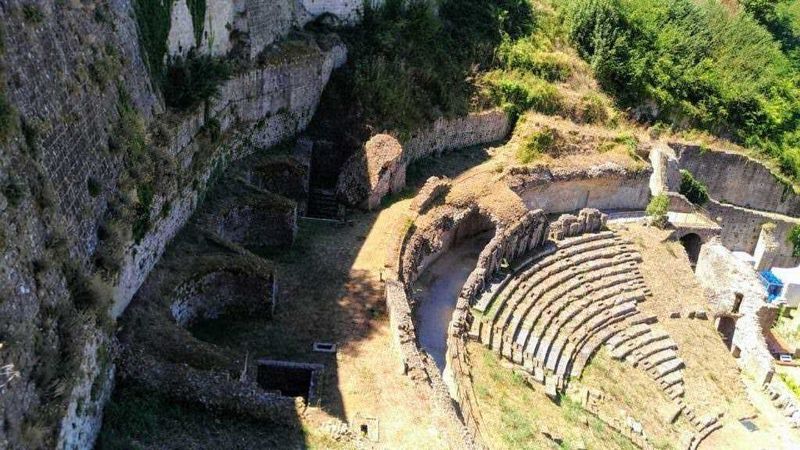 Antico teatro romano in pietra con gradinate e strutture storiche immerso nel verde, esempio di archeologia romana e siti storici italiani.