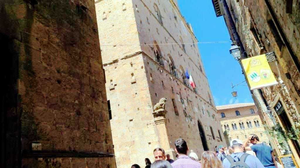 Faro di Torre del Mangia a Siena, Italia, con turisti che ammirano l'architettura storica, cielo azzurro, e atmosfera vivace nel centro storico.