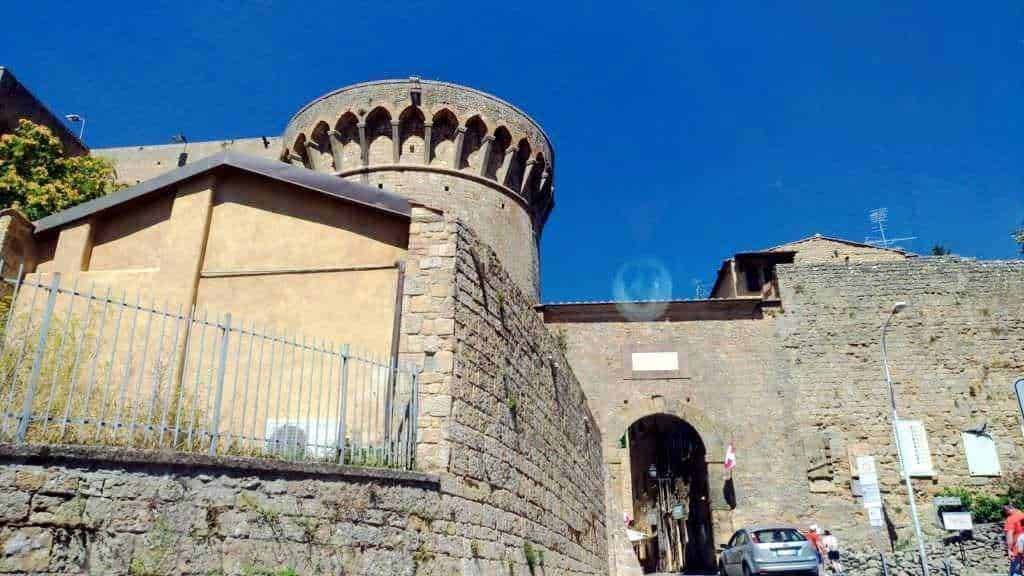 Antico castello medievale con torre circolare in pietra, archi e mura storiche, situato in un borgo italiano, sotto un cielo azzurro, esempio di patrimonio storico e architettonico.
