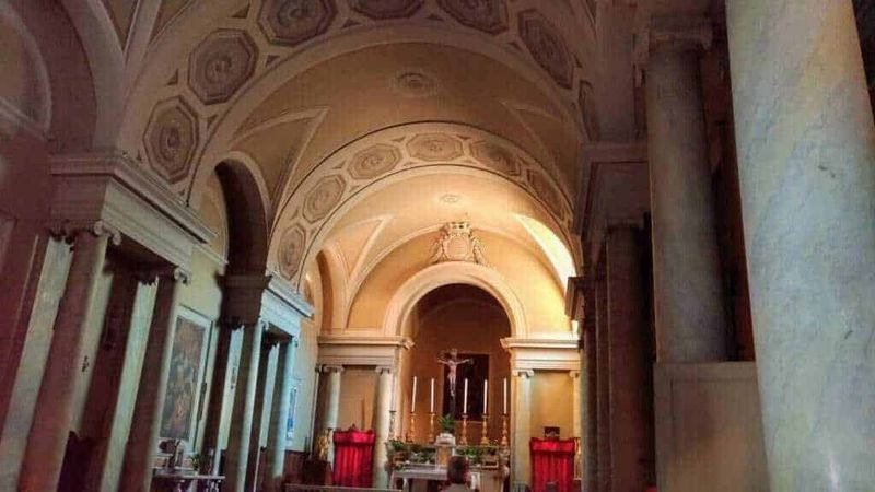 Alt text: Interno chiesa barocca con archi, colonne e altare con crocifisso, illuminata da luce calda, decorazioni in stile classico e dettagli dorati, atmosfera sacra e suggestiva.