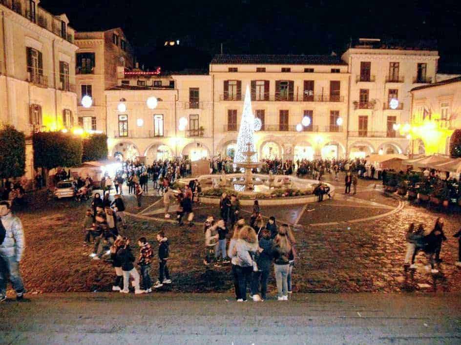 Splendida piazza notturna decorata con luci natalizie e fontana centrale, affollata di persone che passeggiano e si godono l'atmosfera festosa, scenario ideale per scatti di viaggio e turismo culturale.