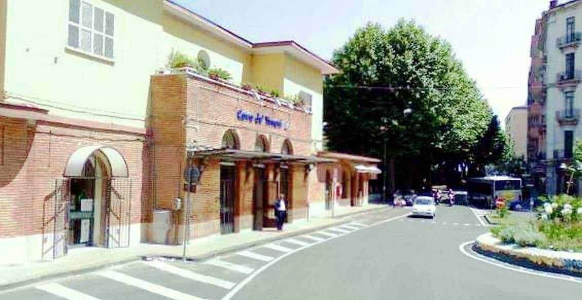 Ingresso di un edificio storico con archi in mattoni rossi in una strada cittadina soleggiata, alberi verdi e veicoli parcheggiati, atmosfera urbana e tranquilla, immagine di un angolo di città, perfetta per articoli sul turismo e la cultura urbana.