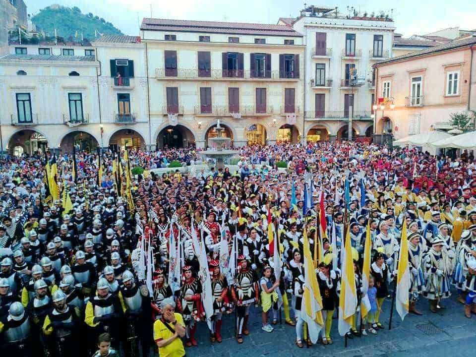 Militari in costume storico in una piazza affollata durante una rievocazione o sagra popolare, con edifici antichi sullo sfondo e moltissime persone partecipanti.