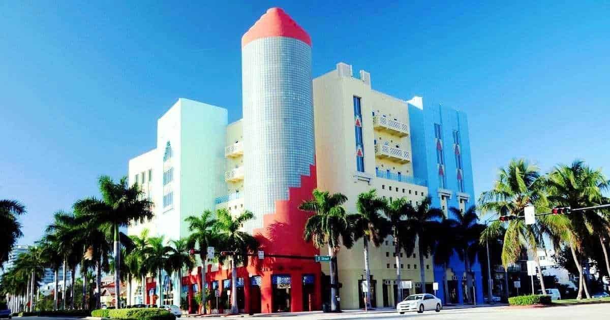 Colorato edificio stile art deco a Miami con palme lungo la strada, cielo azzurro e auto in movimento, ideale per immagini di viaggi, design urbano e destinazioni turistiche.