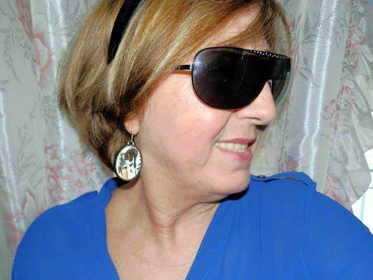 Orecchini fatti a mano in carta riciclata: Elegante donna sorridente con occhiali da sole neri, orecchini pendenti con design originale e abbigliamento blu, in una scena luminosa e raffinata.