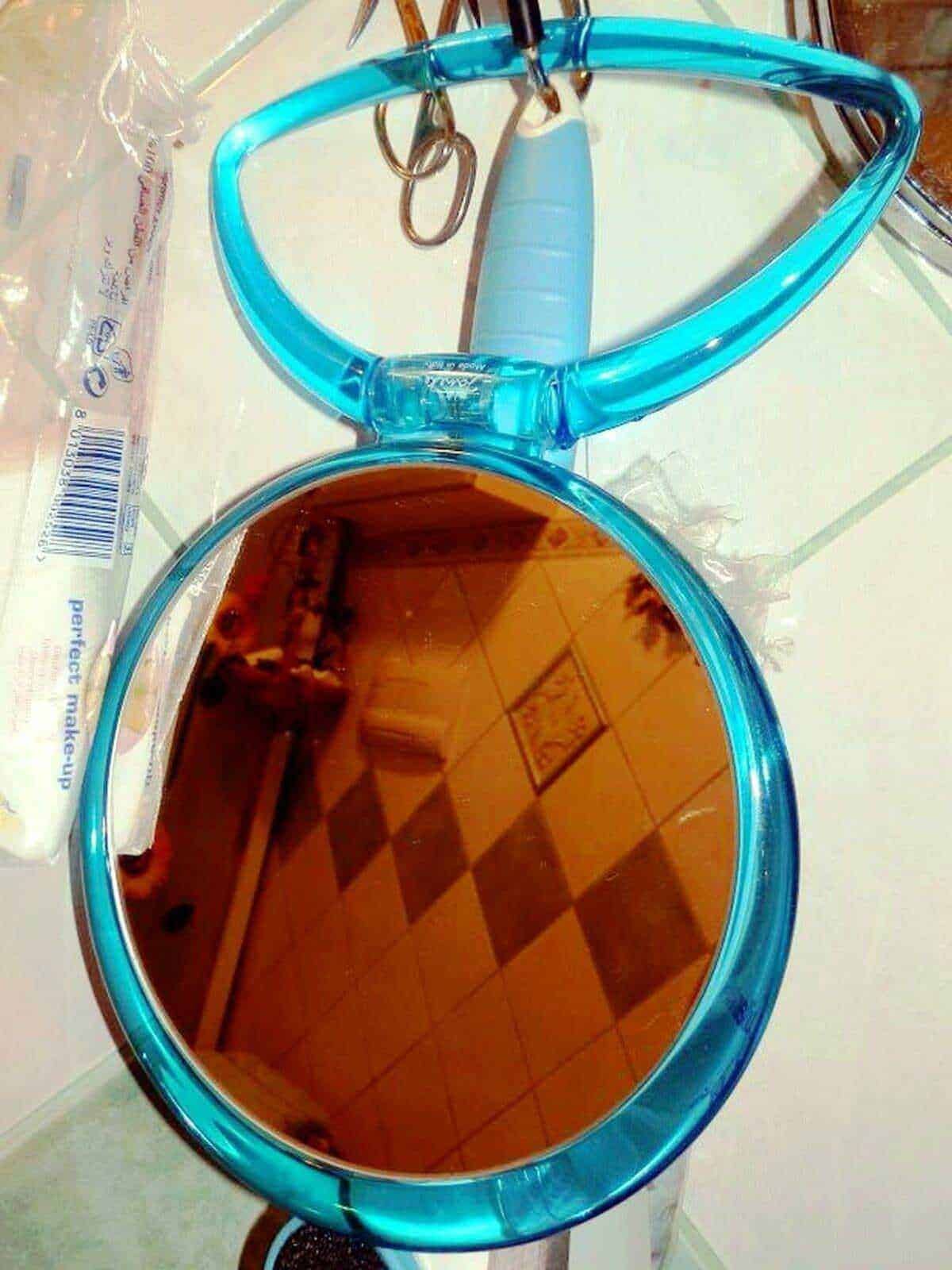 Specchio da trucco con cornice blu trasparente, riflesso sulla superficie mostra un pavimento a quadretti e decorazioni murali. Perfetto per look quotidiani e attenzione ai dettagli di bellezza.