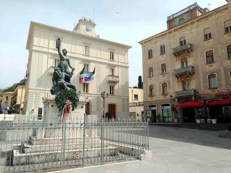 Statuetta commemorativa in piazza con edificio storico sullo sfondo, decorata con corona floreale e bandiere italiane, ambientazione urbana con edifici antichi e caffetterie all'aperto.